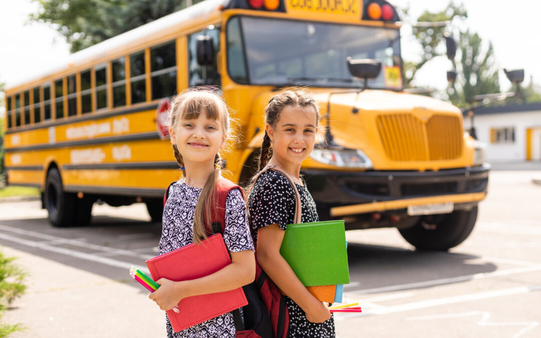 Two young girls stand in front of their school bus, excited for the start of a new school year.