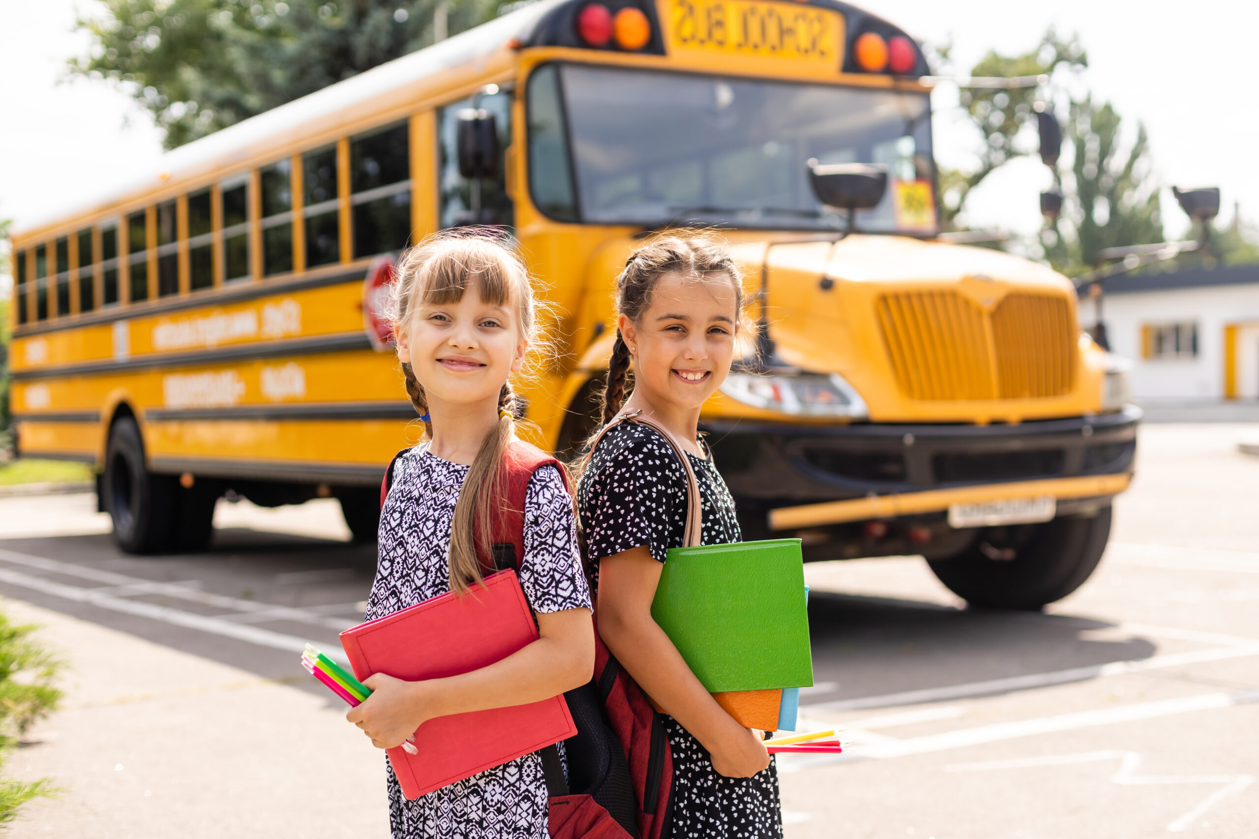 Two young girls stand in front of their school bus, excited for the start of a new school year.